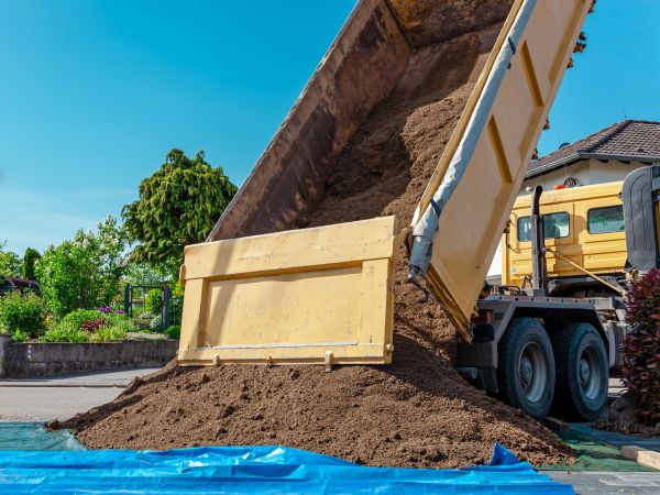 Dump truck tipping a full load of rich brown topsoil onto a tarp in a residential driveway